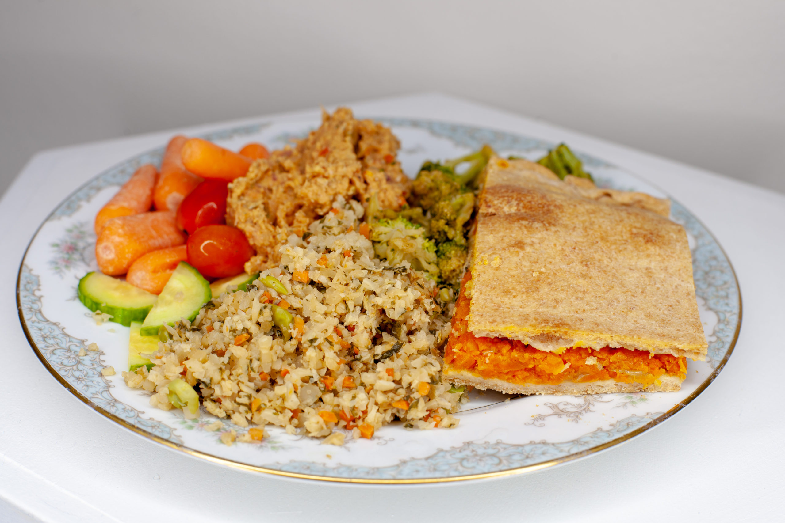 A piece of savoury carrot pie served with various foods, cauliflower fried rice, broccoli stir fry, fresh baby carrots and cherry tomatoes with bean pate dip